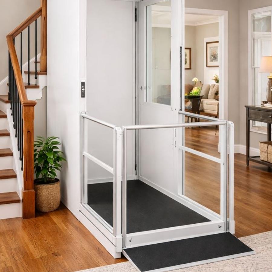 A white indoor vertical platform lift with a glass door and non-slip ramp installed next to a staircase in a modern home living room.