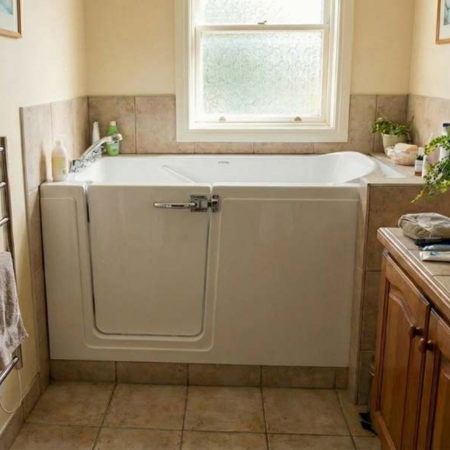 Hilton Head SC walk-in tub in a compact beige tiled bathroom with side-entry door, chrome faucet, safety features and nearby wooden vanity for accessible senior bathing and aging in place.